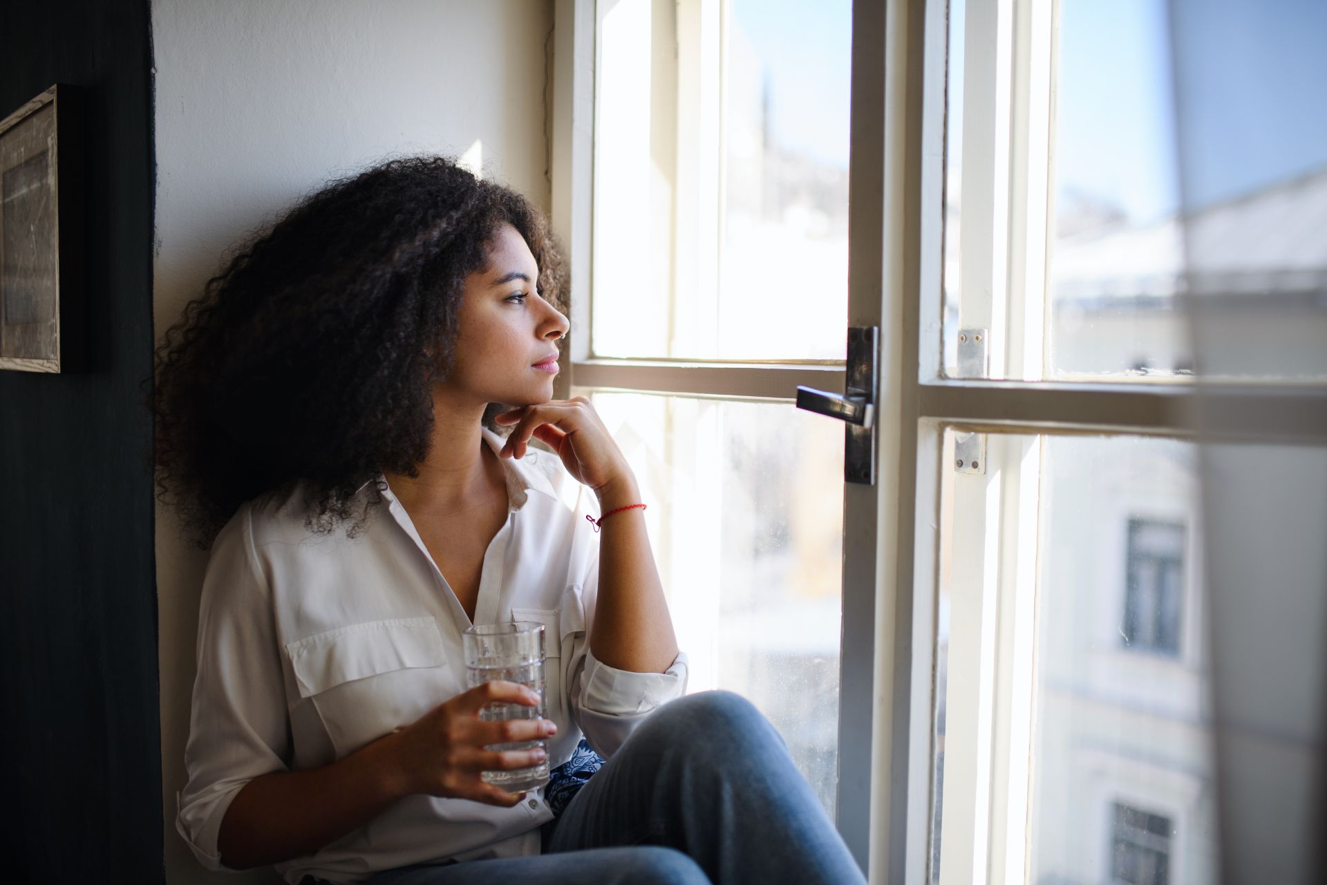 Young mixed race woman sitting on window sill staring into distance with a glass of water in her hand